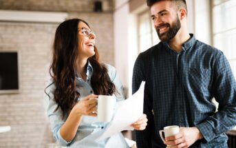 Two people are standing and smiling at each other in an office setting. One holds a coffee mug and papers, with long hair and glasses, while the other has short hair and holds a mug. Both wear casual shirts, and there is a bright, open atmosphere.