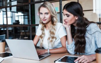 Two women sit at a table with a laptop and a tablet. One woman, in a white shirt with long blonde hair, works on the computer. The other, with long brown hair and a gray top, looks on thoughtfully. Coffee cups and a notebook are also on the table.