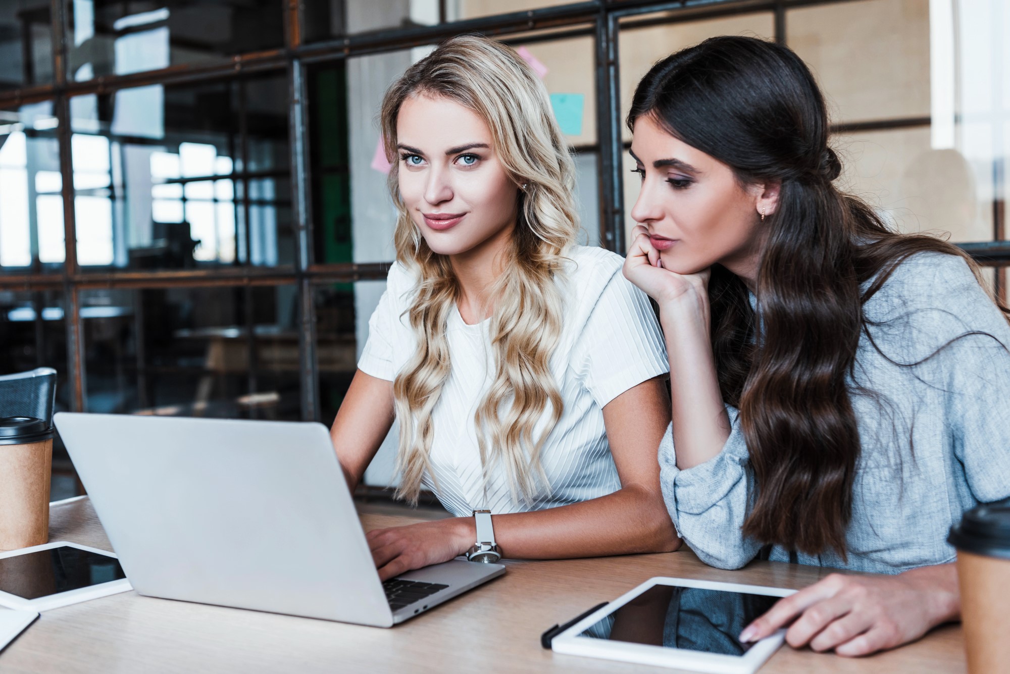 Two women sit at a table with a laptop and a tablet. One woman, in a white shirt with long blonde hair, works on the computer. The other, with long brown hair and a gray top, looks on thoughtfully. Coffee cups and a notebook are also on the table.