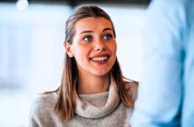 A woman with long hair smiles and looks up attentively. She wears a light-colored turtleneck sweater. The background is softly blurred, suggesting an indoor setting with natural light.