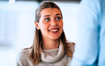 A woman with long hair smiles and looks up attentively. She wears a light-colored turtleneck sweater. The background is softly blurred, suggesting an indoor setting with natural light.