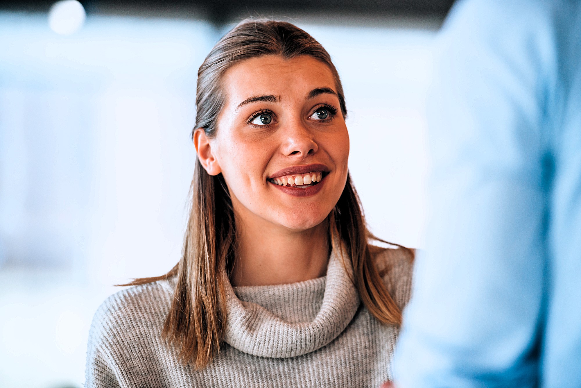 A woman with long hair smiles and looks up attentively. She wears a light-colored turtleneck sweater. The background is softly blurred, suggesting an indoor setting with natural light.