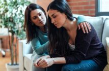 A woman with long dark hair sits on a couch looking pensive, while another woman with similar hair rests her hand on her shoulder in comfort. They're in a cozy living room with a plant and a large window in the background.