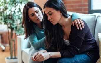 A woman with long dark hair sits on a couch looking pensive, while another woman with similar hair rests her hand on her shoulder in comfort. They're in a cozy living room with a plant and a large window in the background.