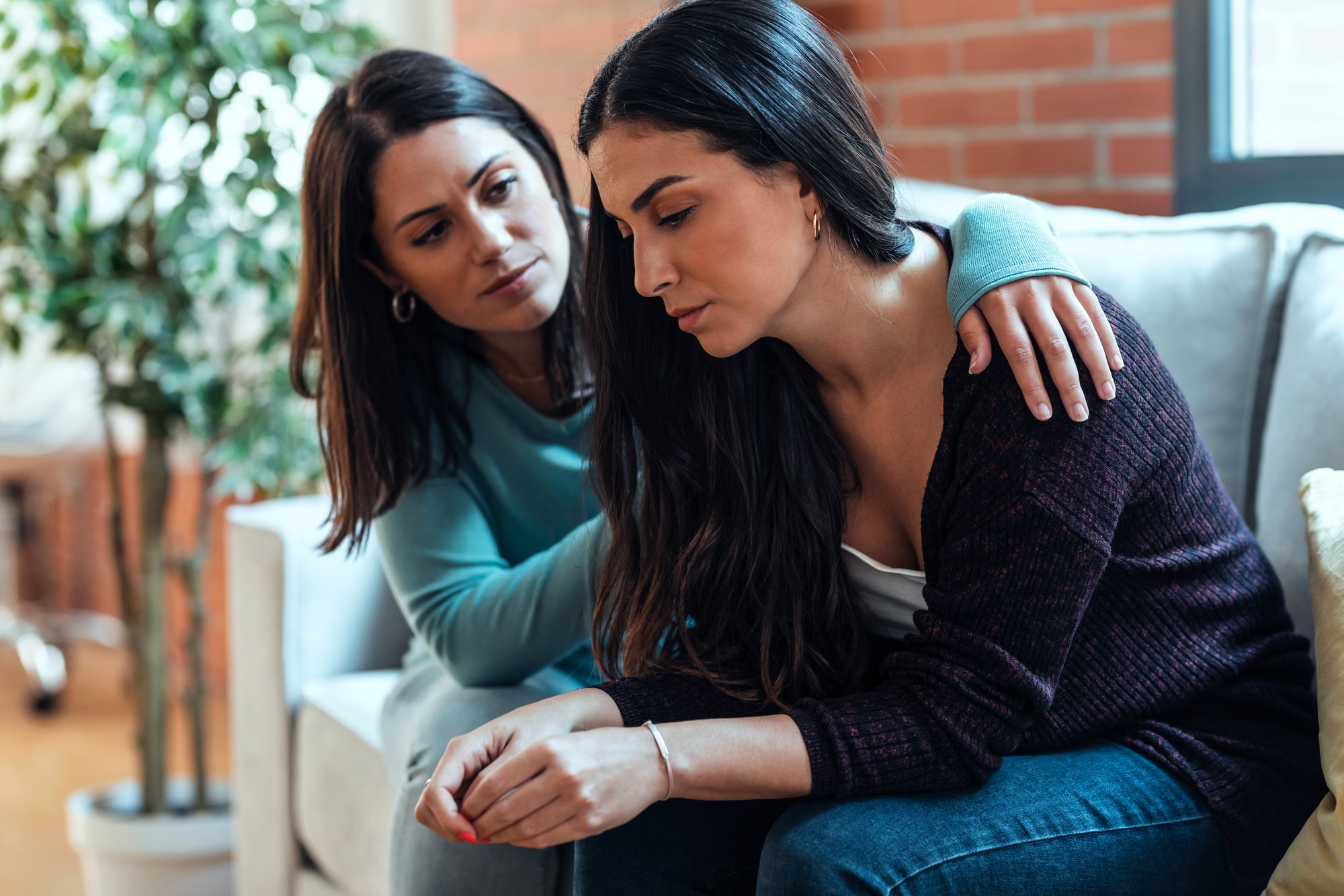 A woman with long dark hair sits on a couch looking pensive, while another woman with similar hair rests her hand on her shoulder in comfort. They're in a cozy living room with a plant and a large window in the background.