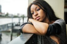 A woman with long brown hair leans on a railing, resting her head on her arms and looking thoughtfully into the distance. She is wearing a gray and black top, and a blurred waterside cityscape is visible in the background.