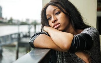 A woman with long brown hair leans on a railing, resting her head on her arms and looking thoughtfully into the distance. She is wearing a gray and black top, and a blurred waterside cityscape is visible in the background.