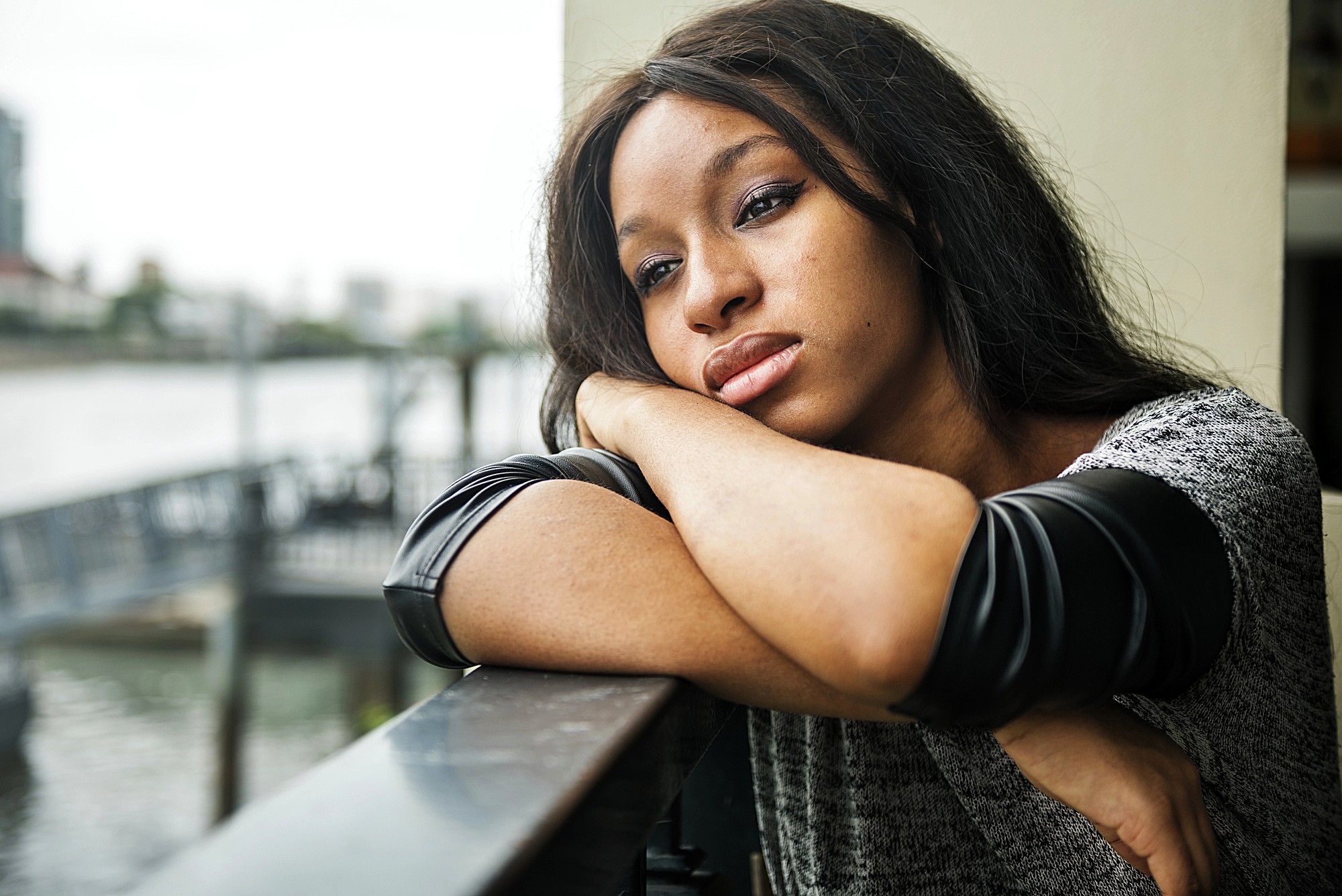 A woman with long brown hair leans on a railing, resting her head on her arms and looking thoughtfully into the distance. She is wearing a gray and black top, and a blurred waterside cityscape is visible in the background.