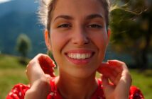 A woman with a nose ring smiles warmly at the camera. She stands outdoors with green hills and a blurred tree in the background. She wears a red patterned top and gently holds her hands near her neck. The sun illuminates her face.