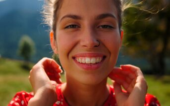 A woman with a nose ring smiles warmly at the camera. She stands outdoors with green hills and a blurred tree in the background. She wears a red patterned top and gently holds her hands near her neck. The sun illuminates her face.