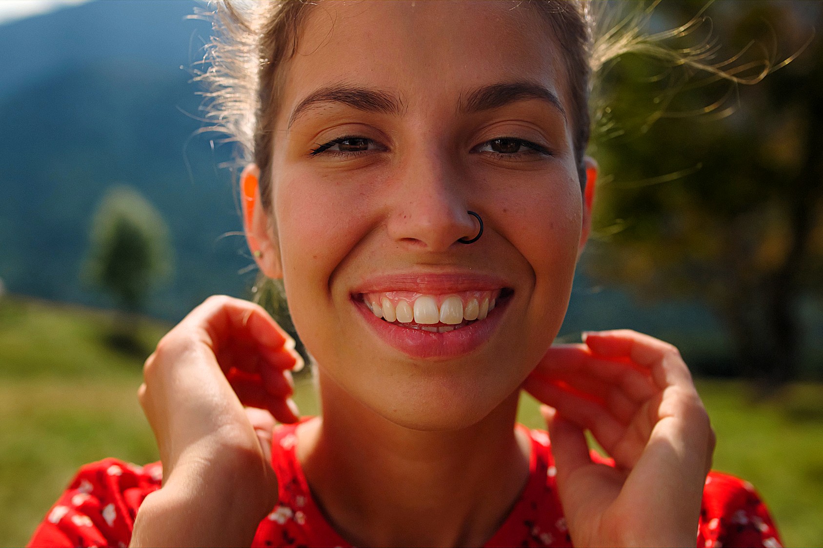 A woman with a nose ring smiles warmly at the camera. She stands outdoors with green hills and a blurred tree in the background. She wears a red patterned top and gently holds her hands near her neck. The sun illuminates her face.