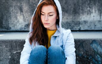 A person with long red hair and freckles sits on concrete steps, wearing a gray hoodie and jeans. They are looking down with a calm expression. The background is a plain, textured concrete wall.