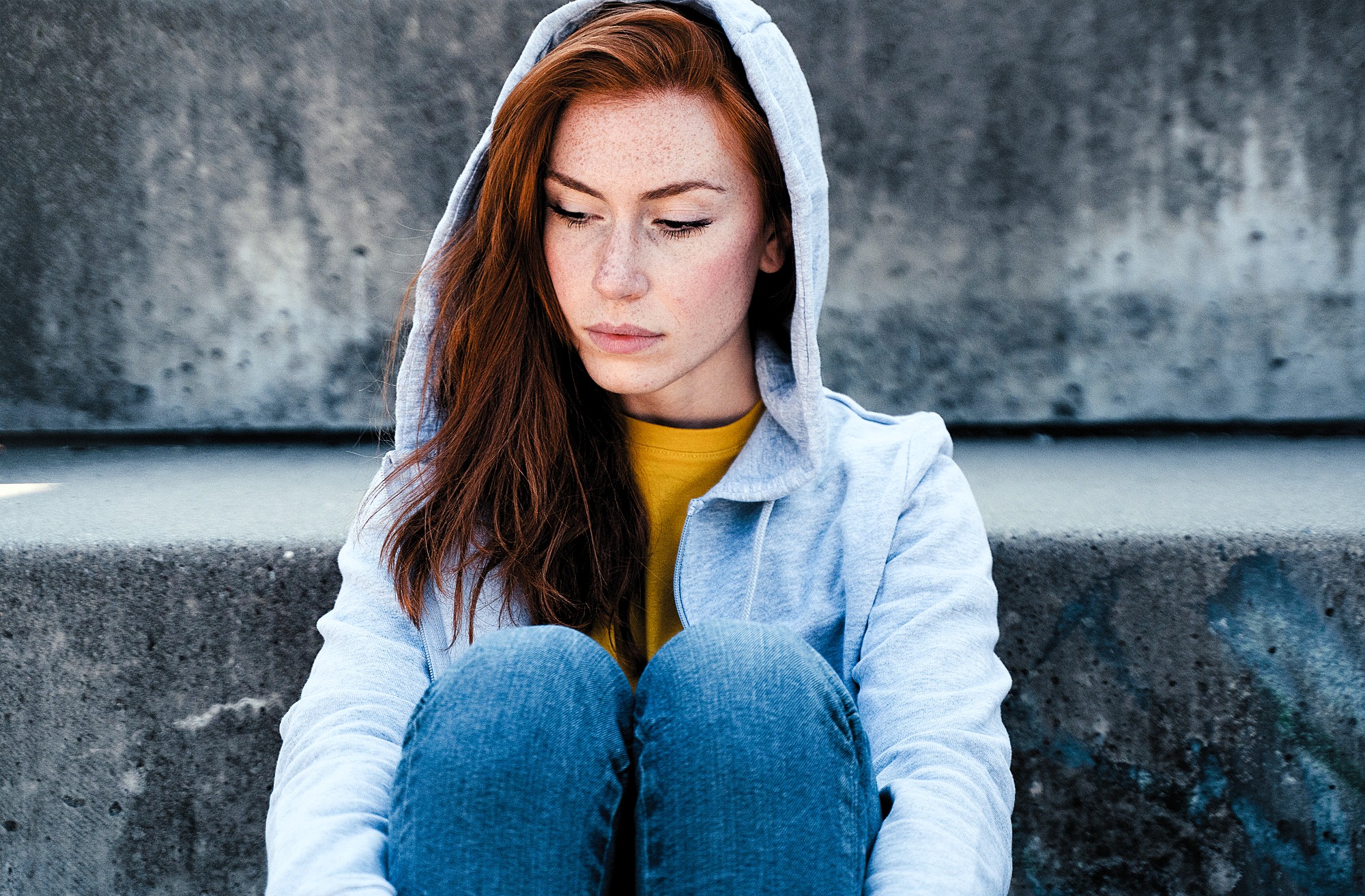 A person with long red hair and freckles sits on concrete steps, wearing a gray hoodie and jeans. They are looking down with a calm expression. The background is a plain, textured concrete wall.