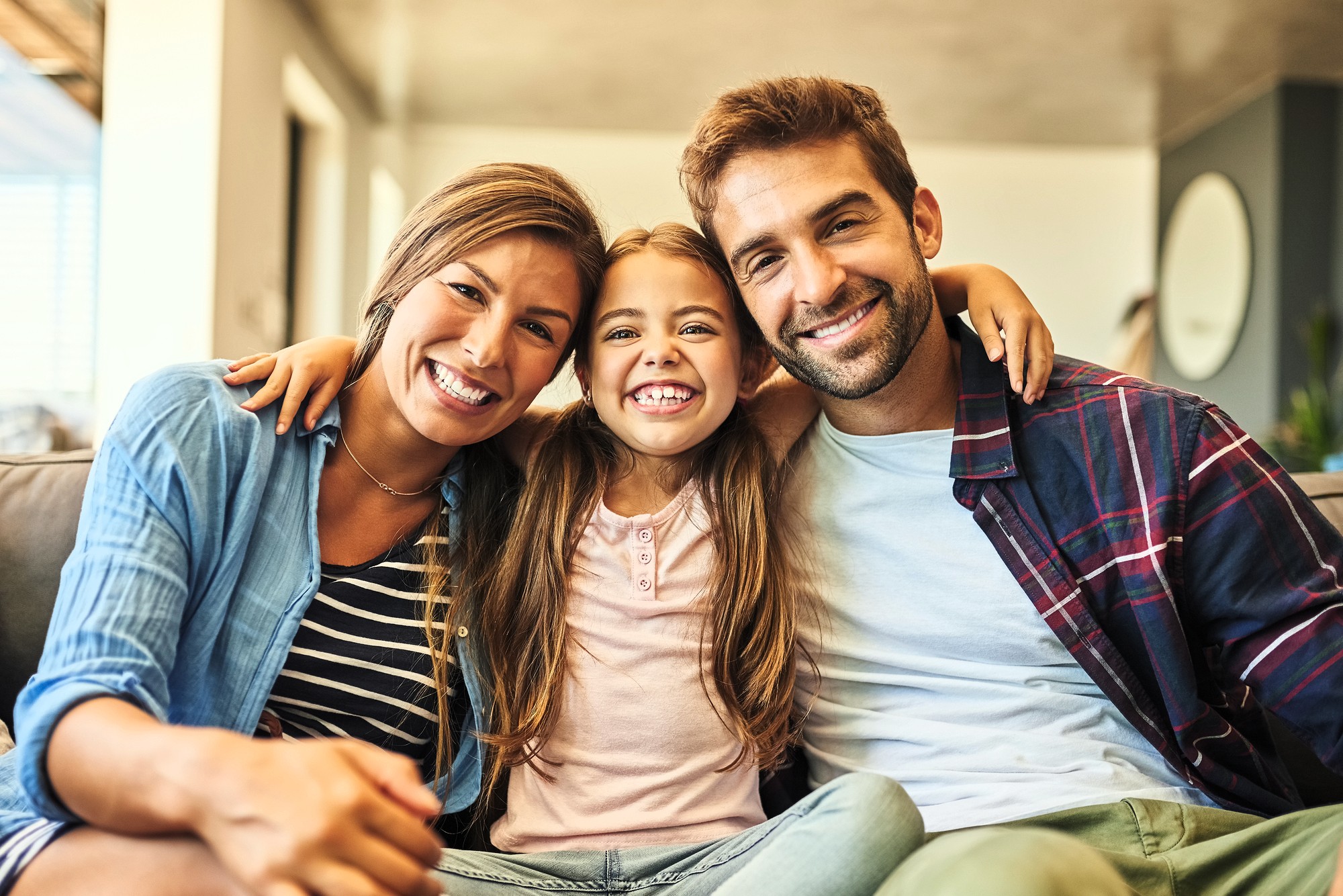 A smiling family of three sits on a couch, with a woman on the left, a young girl in the middle, and a man on the right. The girl has her arms around both adults, and they are all looking at the camera. The setting appears to be a home.