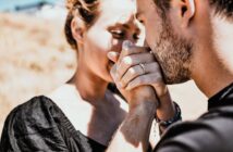 A couple sharing an intimate moment outdoors. The man is gently kissing the woman's hand, which has an engagement ring. They are dressed in casual black attire, with natural sunlight highlighting their expressions. The background is blurred.