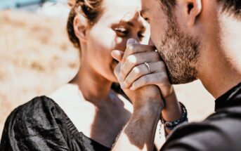 A couple sharing an intimate moment outdoors. The man is gently kissing the woman's hand, which has an engagement ring. They are dressed in casual black attire, with natural sunlight highlighting their expressions. The background is blurred.