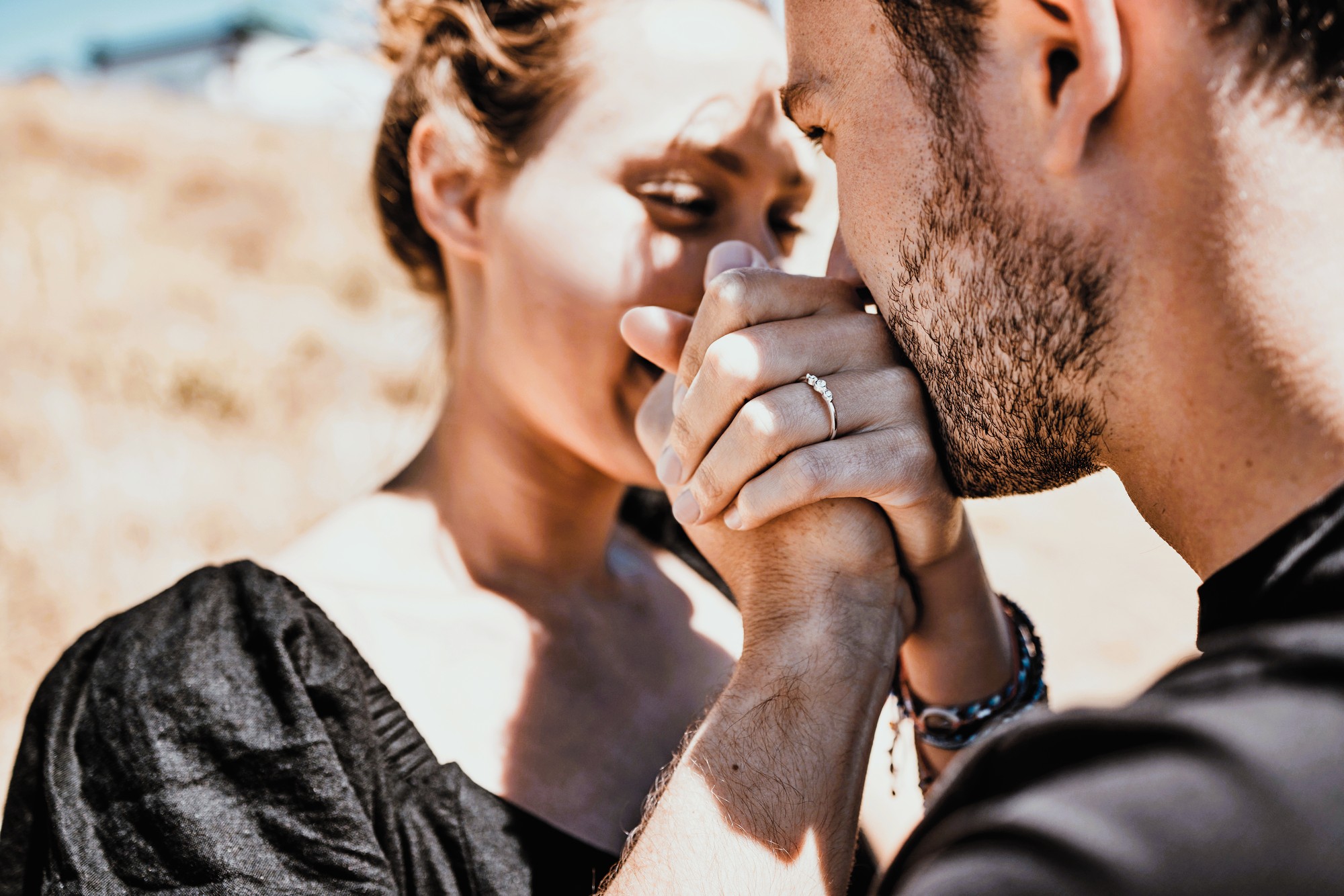 A couple sharing an intimate moment outdoors. The man is gently kissing the woman's hand, which has an engagement ring. They are dressed in casual black attire, with natural sunlight highlighting their expressions. The background is blurred.