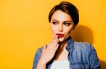 A woman with short brown hair in a denim jacket playfully holds her hand to her lips, looking sideways with a mischievous expression. The background is a bright yellow.