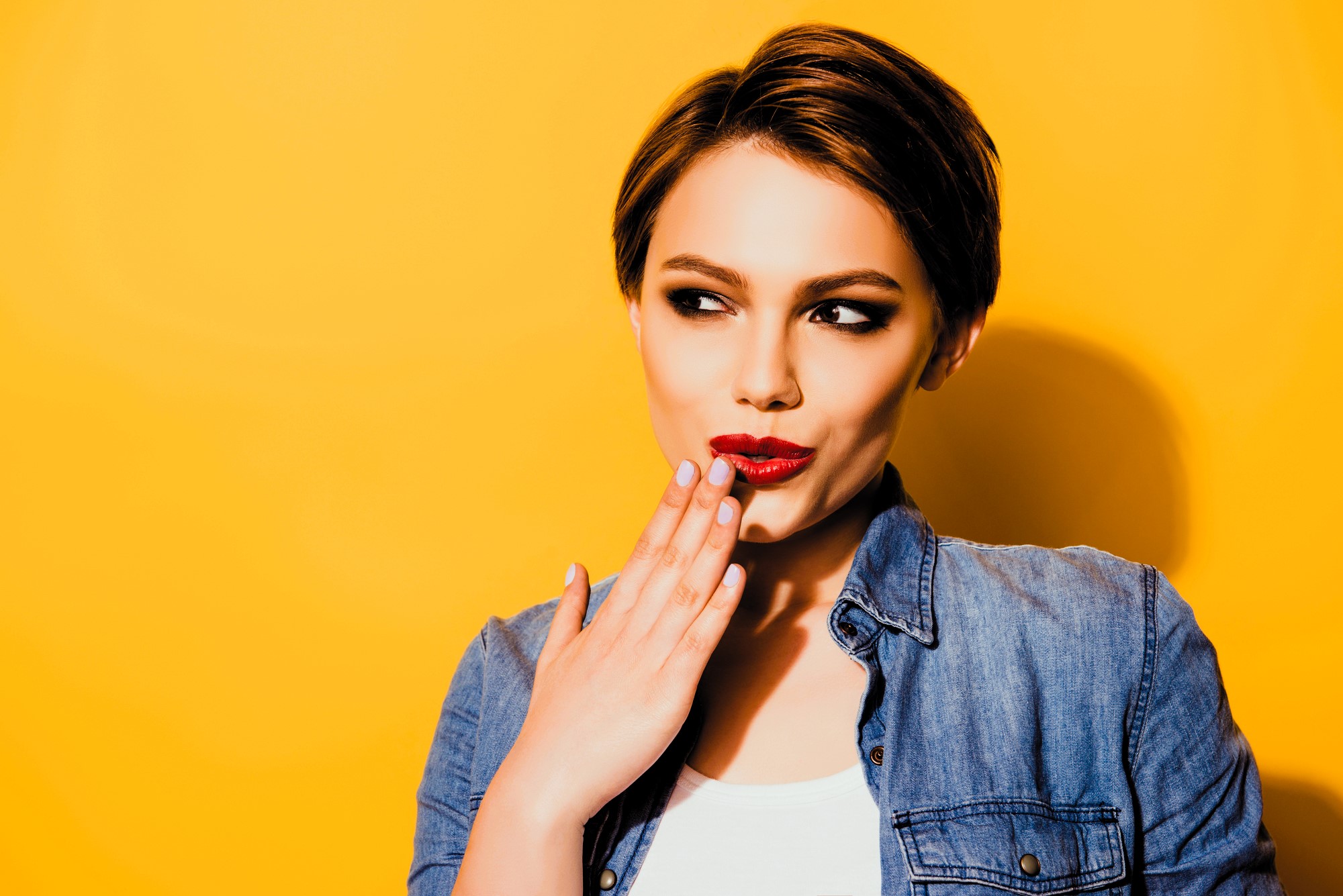 A woman with short brown hair in a denim jacket playfully holds her hand to her lips, looking sideways with a mischievous expression. The background is a bright yellow.