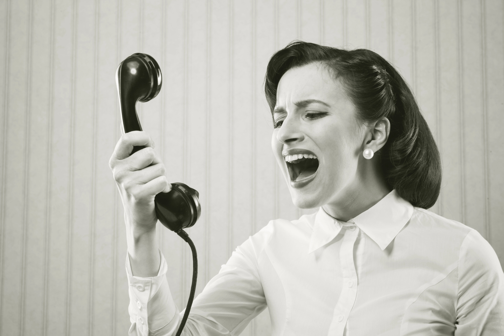 A black-and-white image of a woman in a white blouse, with pearl earrings, holding up a vintage telephone receiver. She appears to be shouting into it, with a frustrated expression. The background has vertical striped wallpaper.