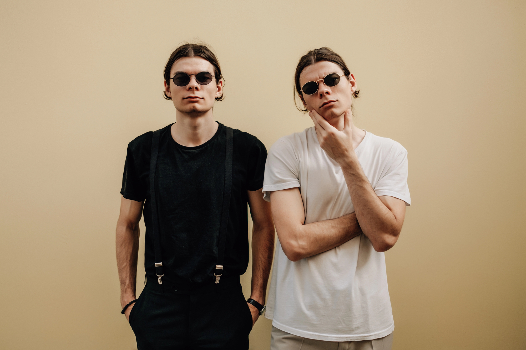 Two people wearing sunglasses stand against a beige background. One wears a black shirt with suspenders and pants, hands in pockets. The other wears a white t-shirt and pants, with one hand on their chin, posing thoughtfully.