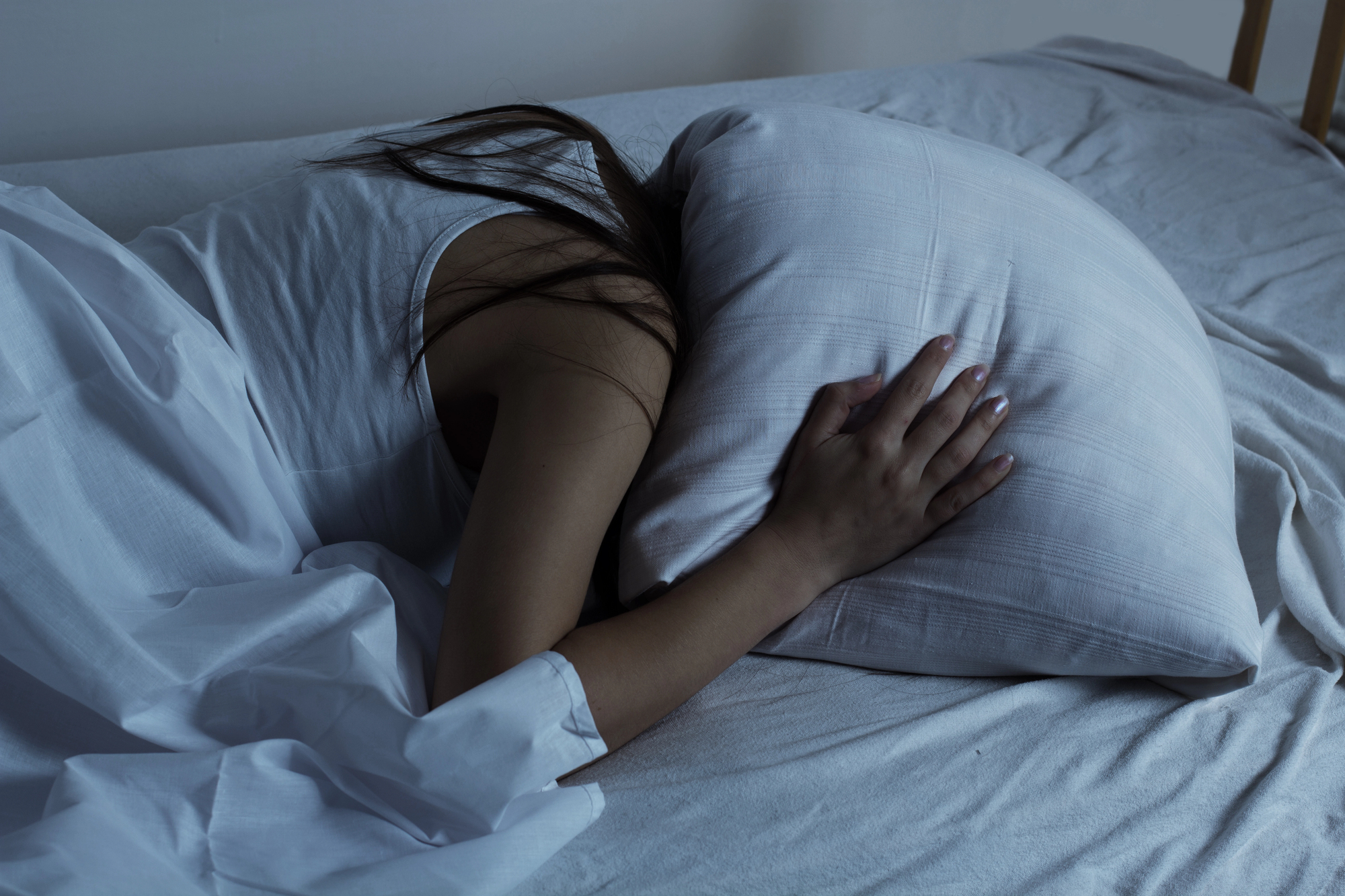 A person lying on a bed with their face buried in a pillow, partially covered by a white sheet. The room is dimly lit, casting soft shadows. The person's arm rests over the pillow, and they are wearing a white tank top.