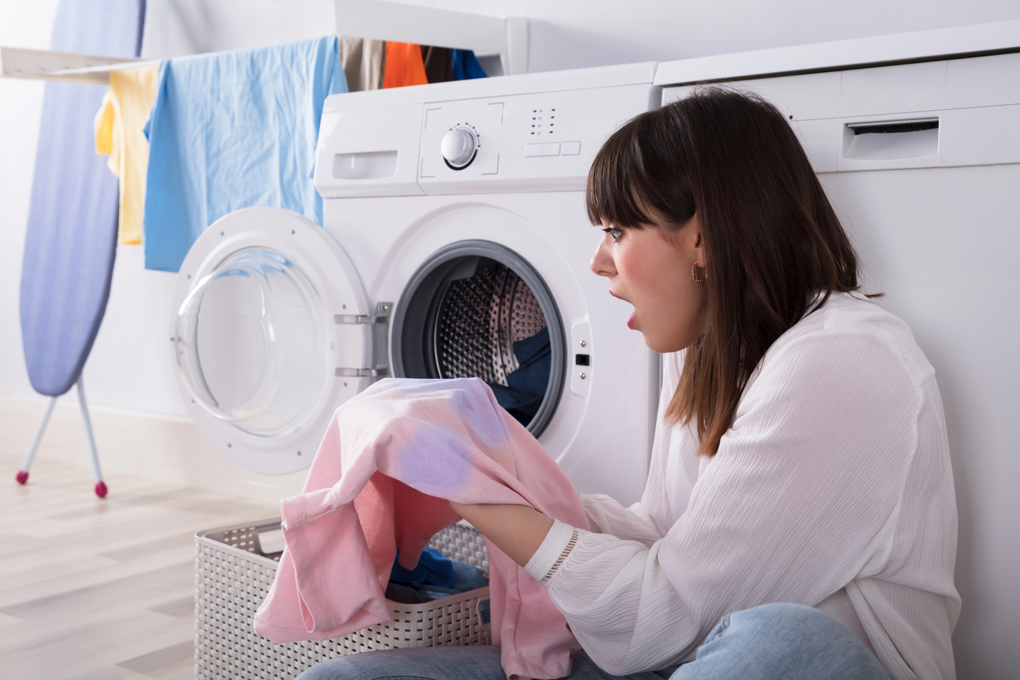 A woman sits on the floor in front of a washing machine, looking surprised at a pink and white garment she's holding. An ironing board and clothes drying on a rack are in the background.