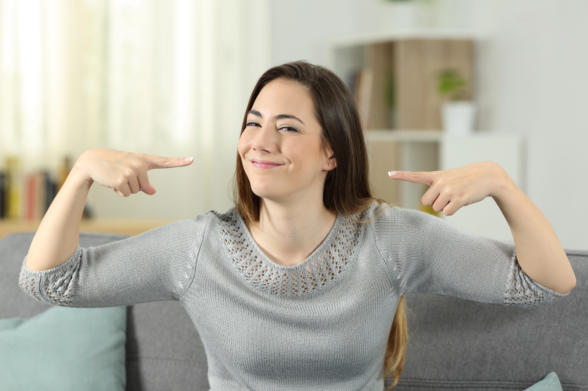 A woman with long brown hair sits on a gray couch, wearing a gray sweater. She is smiling confidently and pointing at herself with both index fingers. The background is softly blurred, showing a bookshelf and a plant.