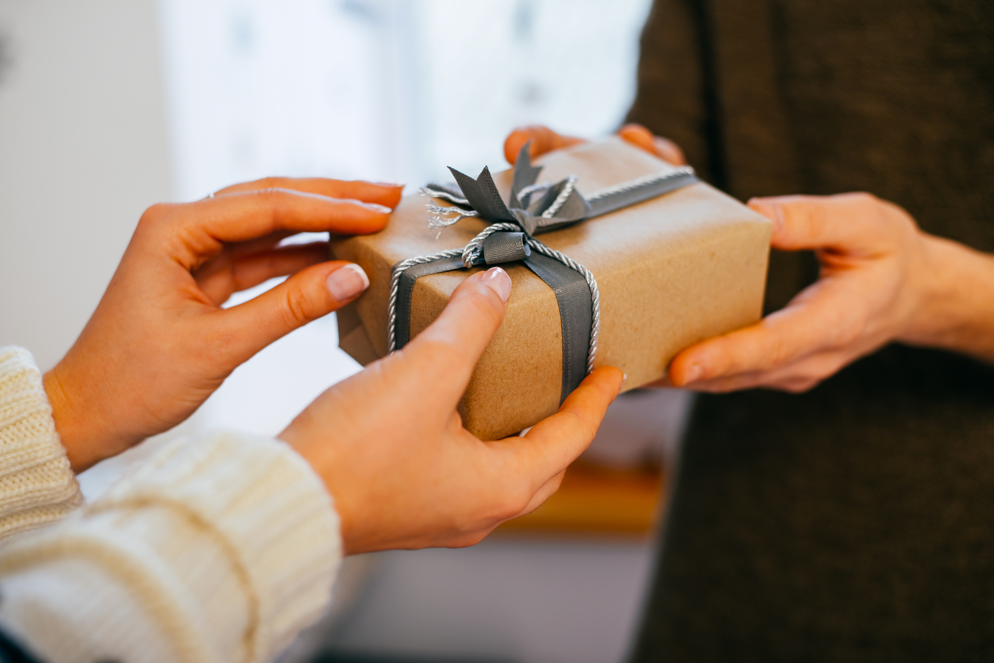 Two people exchanging a gift wrapped in brown paper with a gray ribbon. One person's hands are giving the gift, while the other's are receiving it, evoking a sense of sharing and celebration.