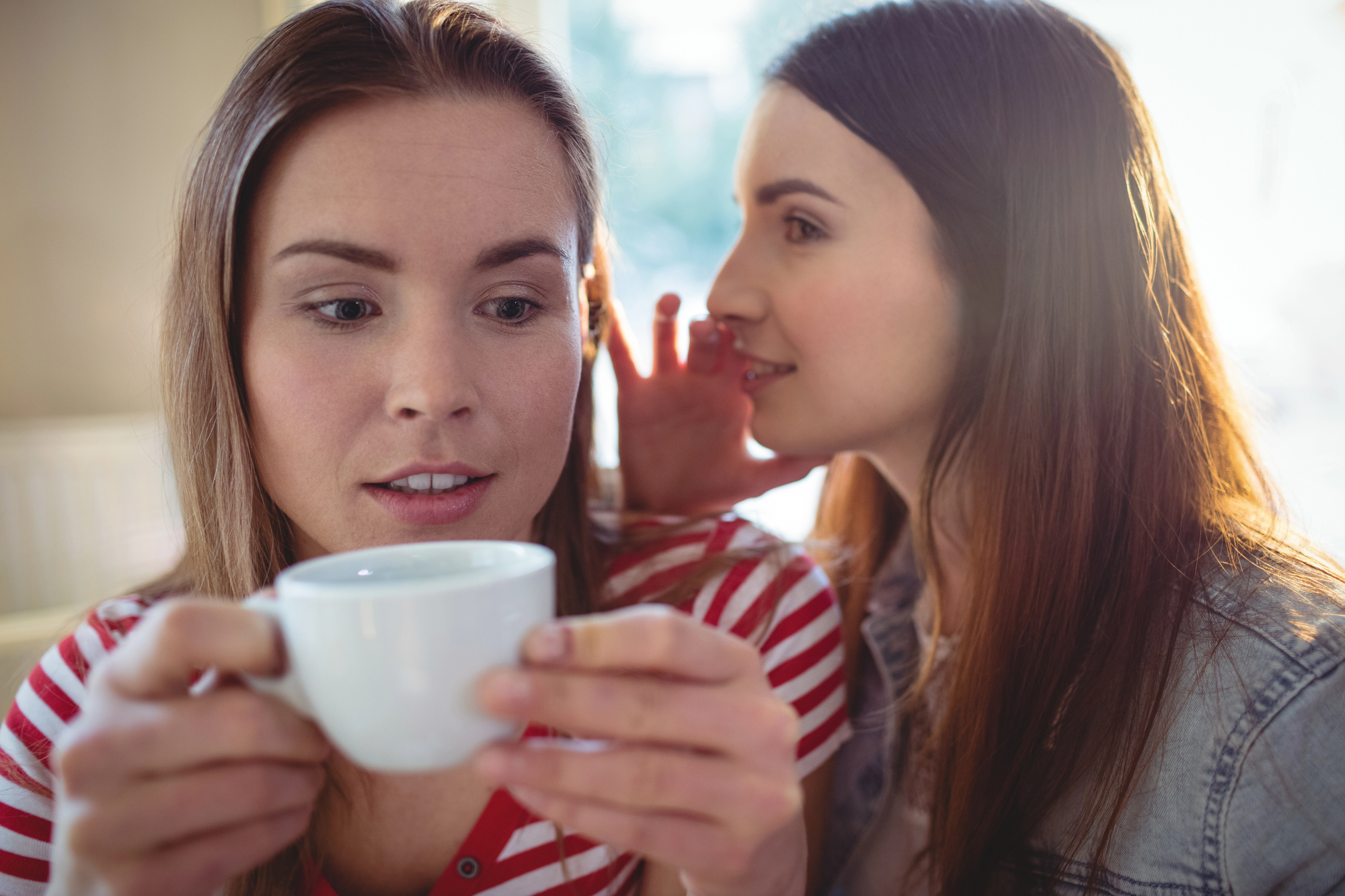 Two women are sitting close together. One woman in a striped shirt is holding a cup, while the other whispers into her ear. The scene appears intimate, with the whispering woman leaning in slightly.