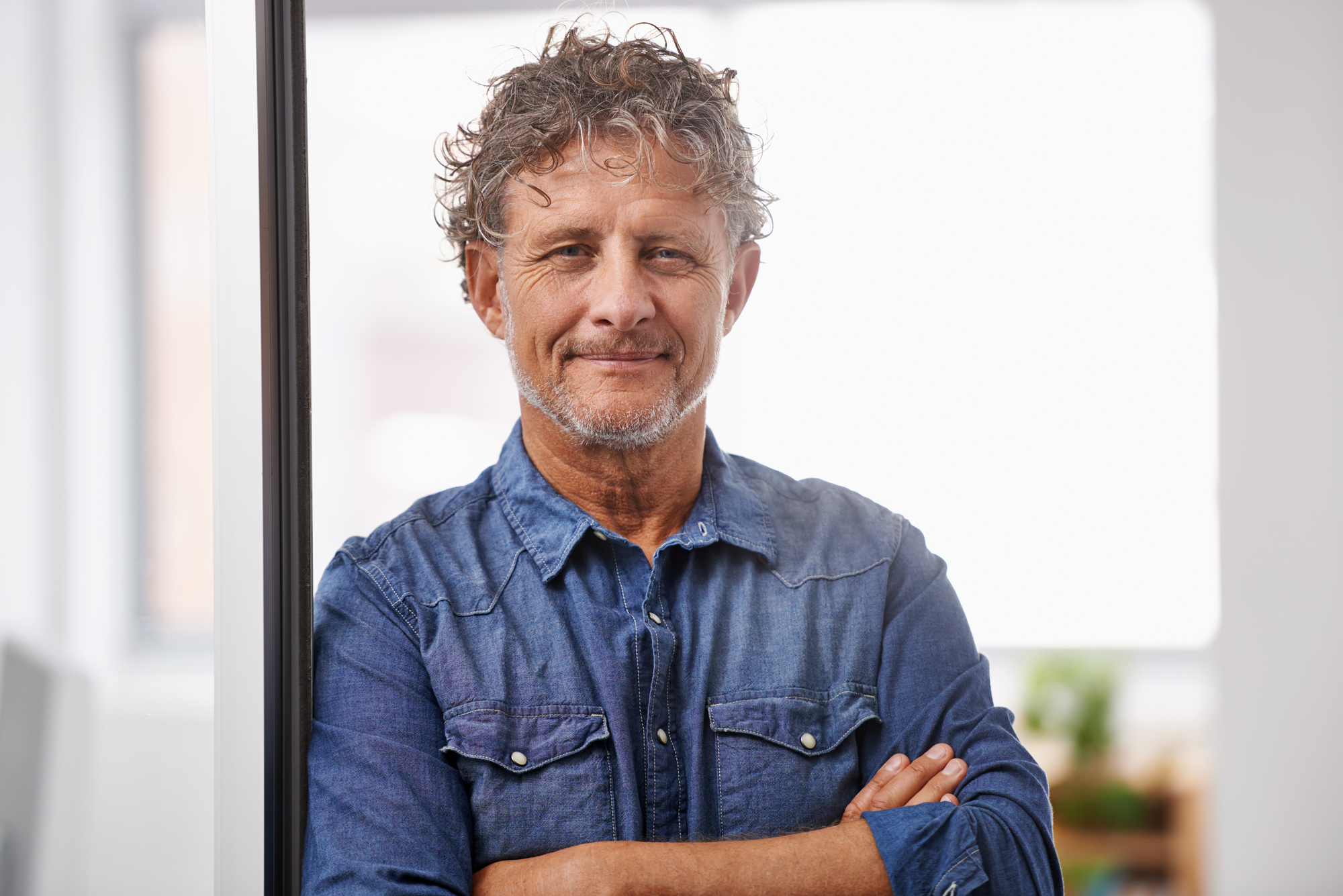 A man with curly hair and a beard stands indoors, wearing a denim shirt. He has his arms crossed and is smiling slightly. The background is softly blurred, suggesting a bright, modern interior.