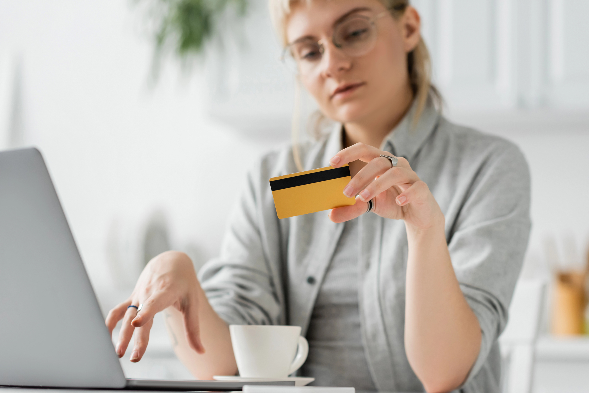 A person sitting at a table holds a gold credit card while looking at a laptop. There is a white cup in front of them. They are wearing glasses and a grey sweater, in a bright room with blurred background.