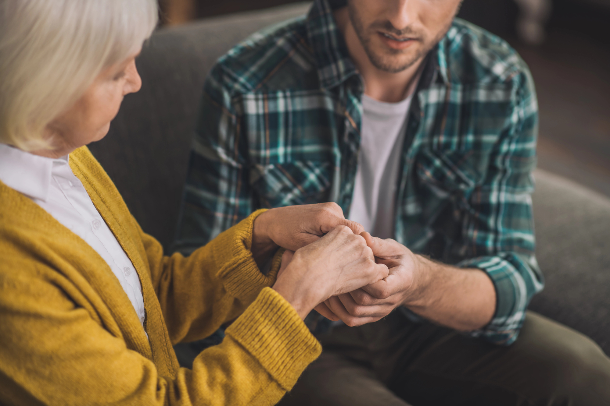 A senior woman in a yellow sweater gently holds the hands of a younger man in a green plaid shirt, sitting on a couch. The scene suggests a comforting or supportive interaction.