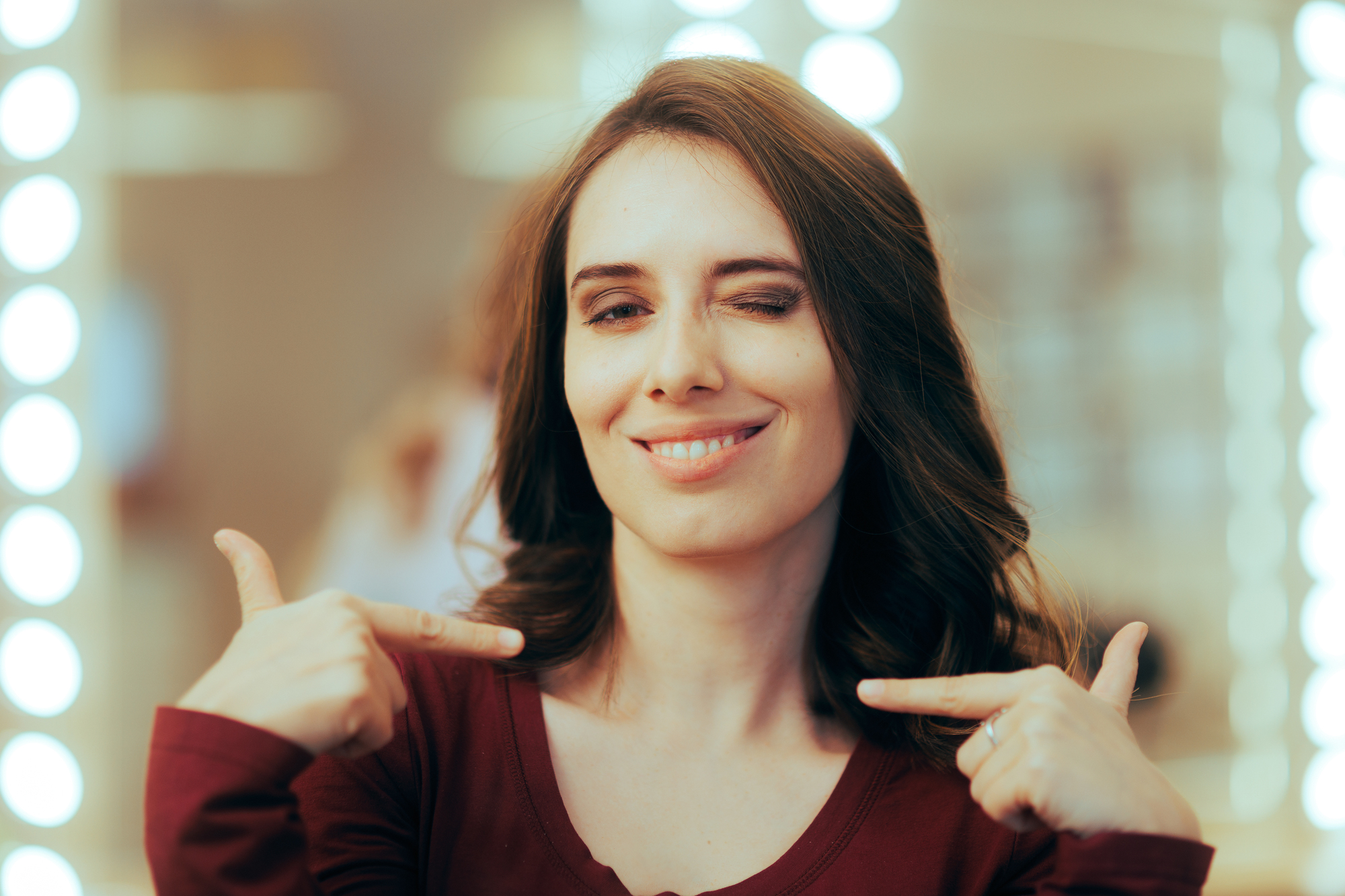 A woman with shoulder-length brown hair is smiling and winking at the camera. She is wearing a maroon top and pointing at herself with both hands. The background is softly blurred with bright circular lights.