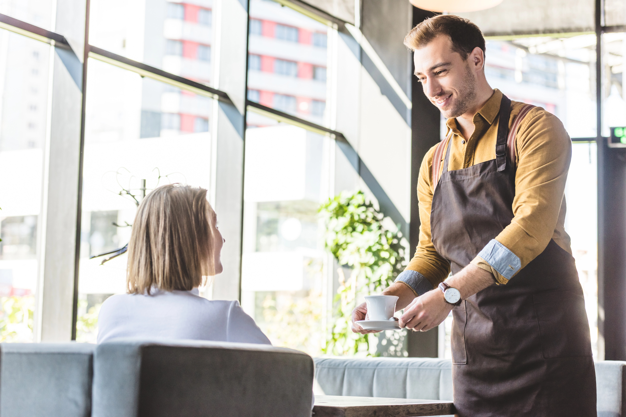 A waiter in a brown apron smiles while serving a cup of coffee to a seated customer in a cafe with large windows. The customer, a woman with shoulder-length blonde hair, looks up at him. Bright daylight filters through the windows.