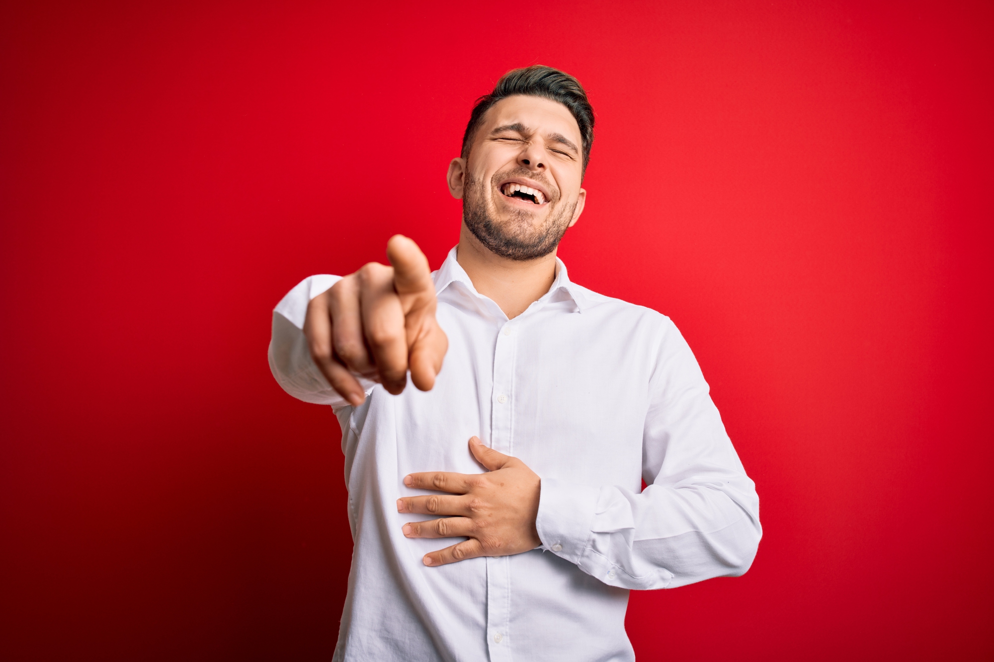 A man in a white shirt stands against a red background, laughing with eyes closed. He points towards the camera with one hand, while the other rests on his stomach.