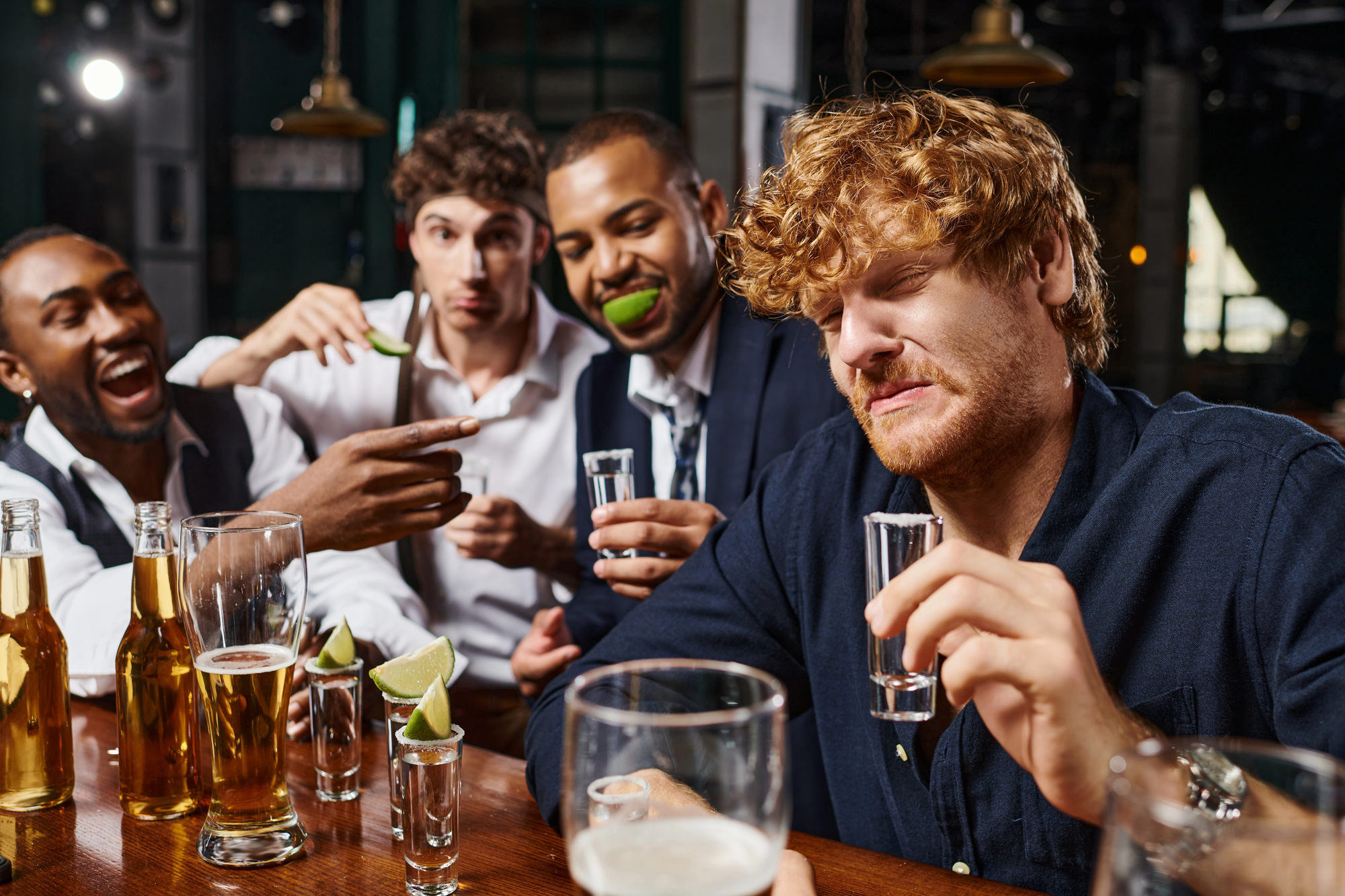 Four men at a bar enjoying drinks. One holds a glass with lime in his mouth, another makes a playful face while holding a shot glass. Others laugh and point, with beer bottles and lime wedges on the table.