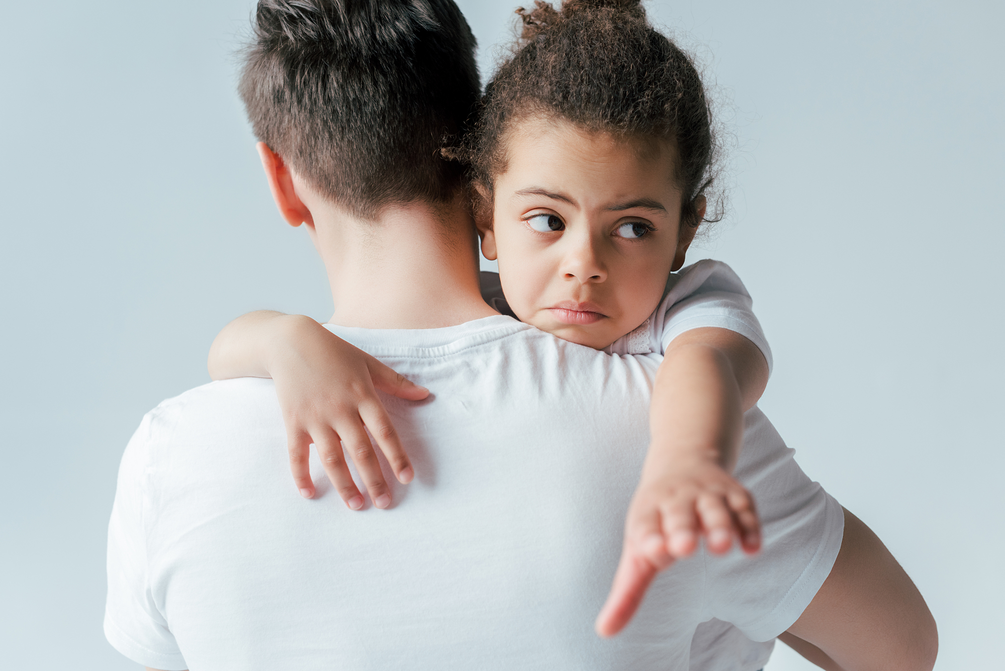 A young child with curly hair, looking concerned, is being held by an adult wearing a white shirt. The child's arm is extended outward. The background is a plain, light color.