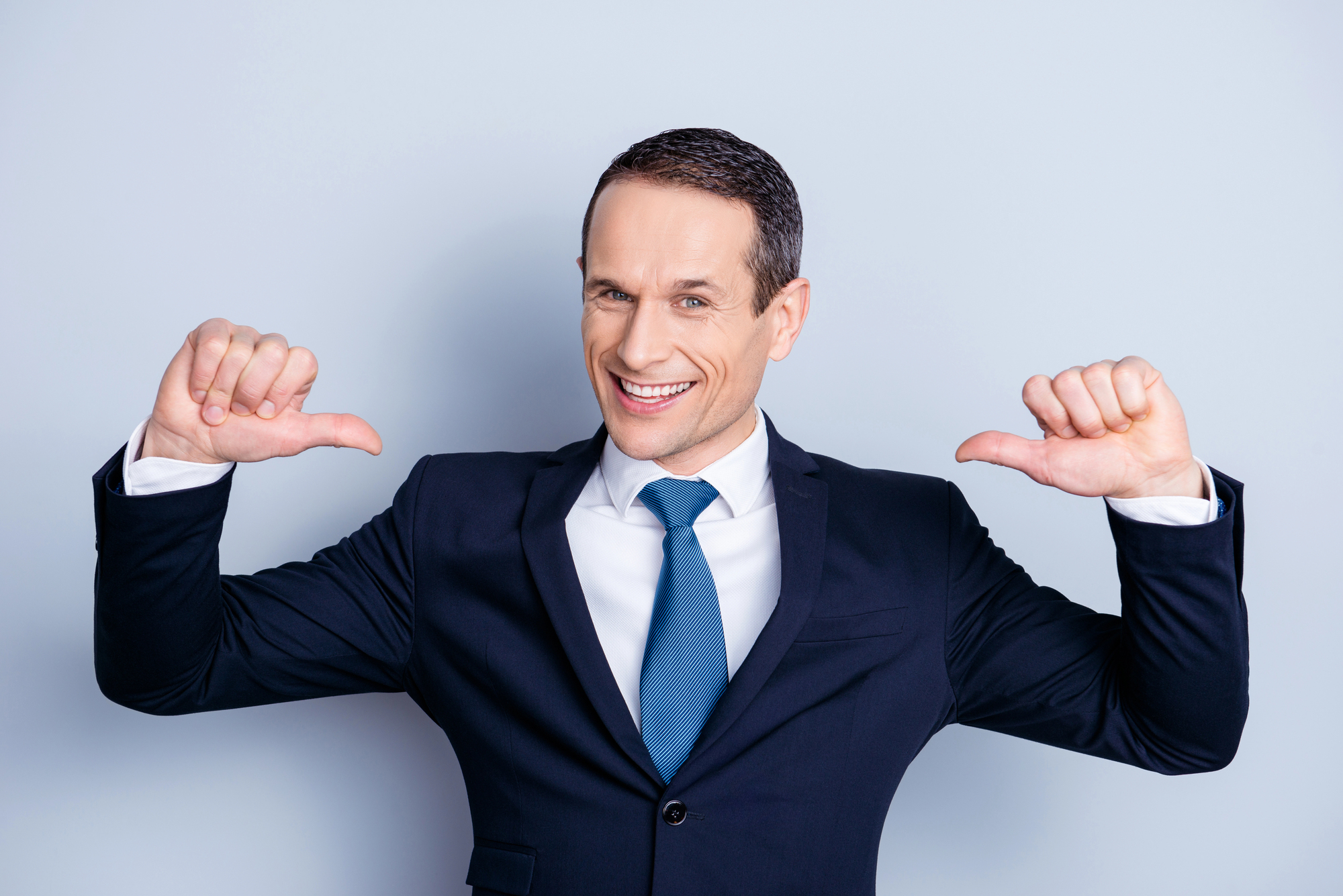 A man in a navy suit and blue tie smiles, pointing his thumbs towards himself. He stands against a plain light blue background.