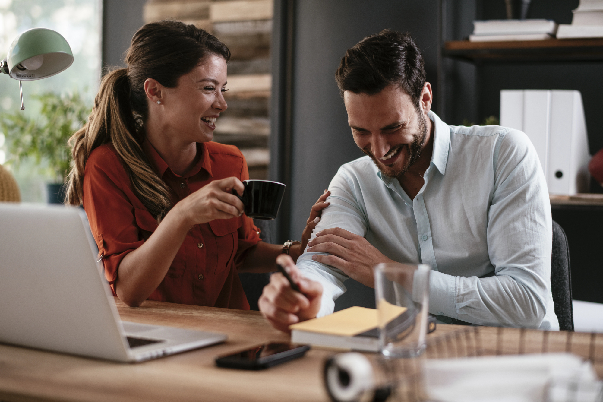 A man and woman are sitting at a desk, laughing and enjoying each other's company. The woman is holding a coffee mug, and the man is writing with a pen. A laptop and a glass of water are visible on the desk.