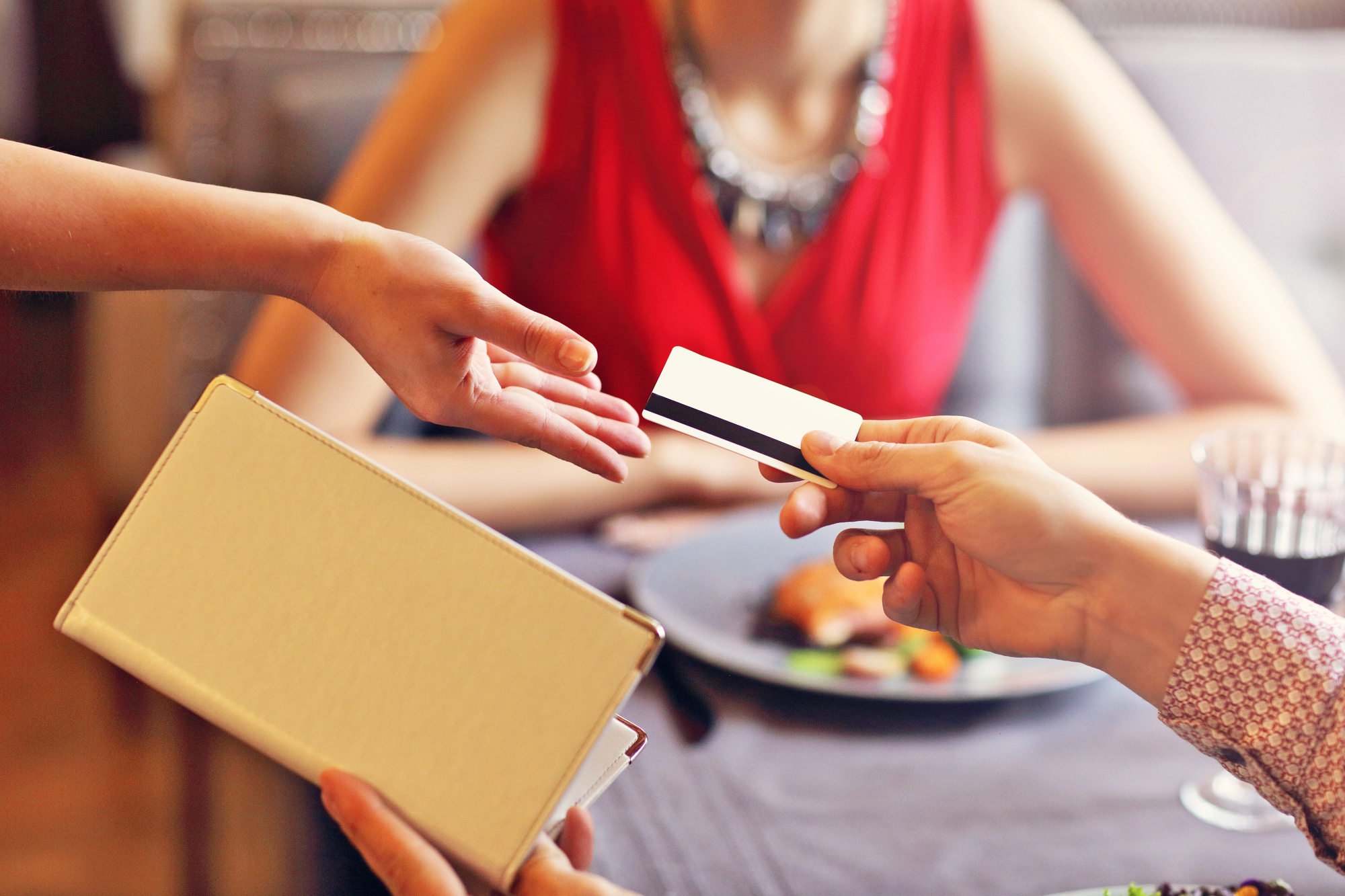 A person hands a credit card to another person's outstretched hand, holding a receipt or bill folder. A third person in a red dress is visible in the background, sitting at a dining table with a plate of food and a glass of wine.