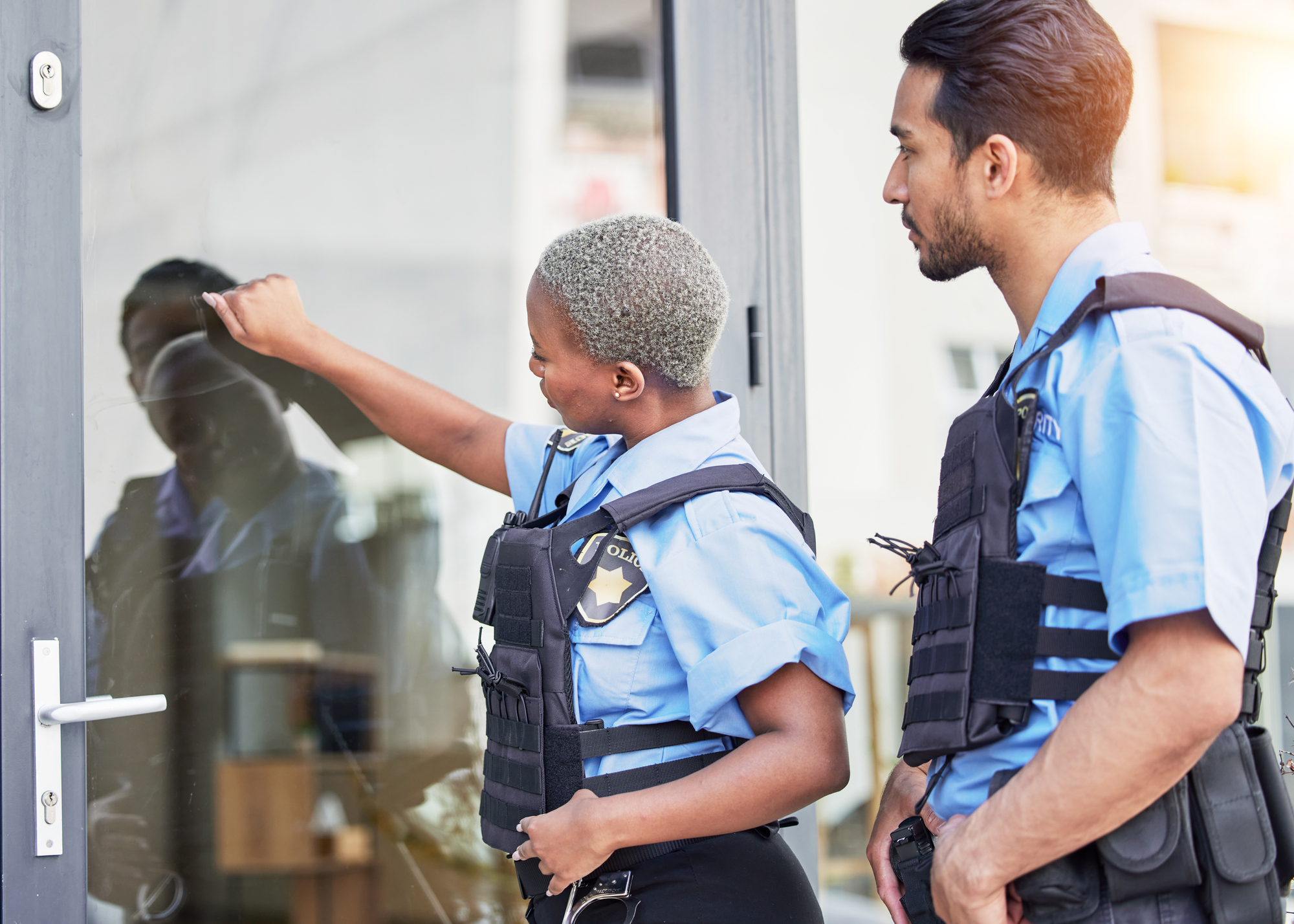 Two security officers in blue uniforms and tactical vests are standing outside a glass door. One is knocking, while the other watches. They are focused on the door, and the reflection of the person knocking is visible in the glass.