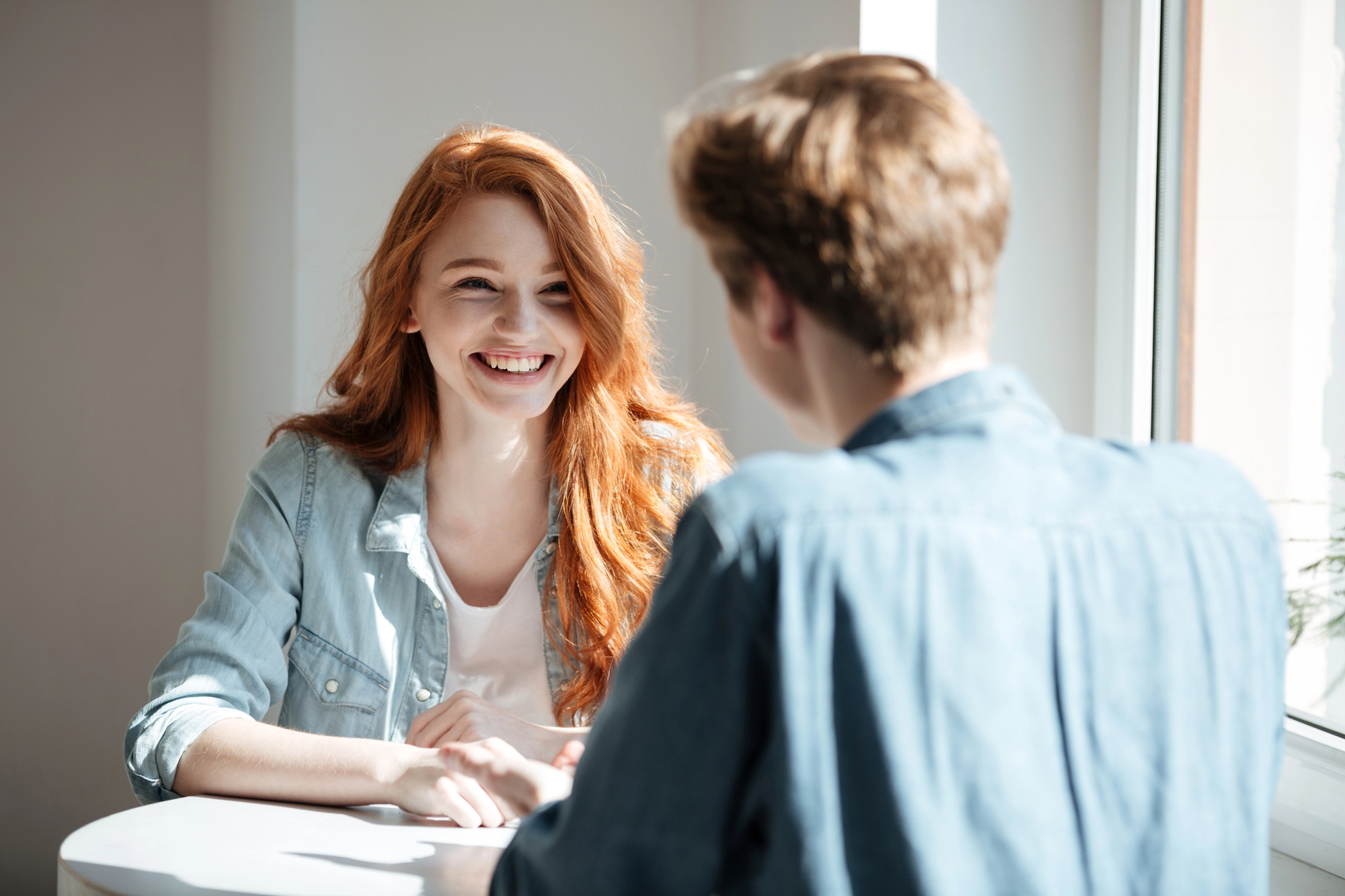 Two people sit across from each other at a round table by a window. The person facing the camera has long red hair and is smiling. Both are wearing casual denim shirts. Sunlight illuminates the scene.