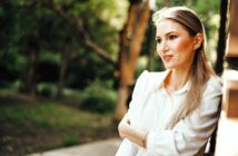 A person with long hair leans against a wall outdoors, wearing a white blouse. They seem thoughtful, looking to the side. The background is green with blurred foliage, suggesting a sunny day in a garden or park setting.