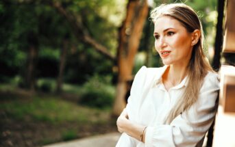 A person with long hair leans against a wall outdoors, wearing a white blouse. They seem thoughtful, looking to the side. The background is green with blurred foliage, suggesting a sunny day in a garden or park setting.