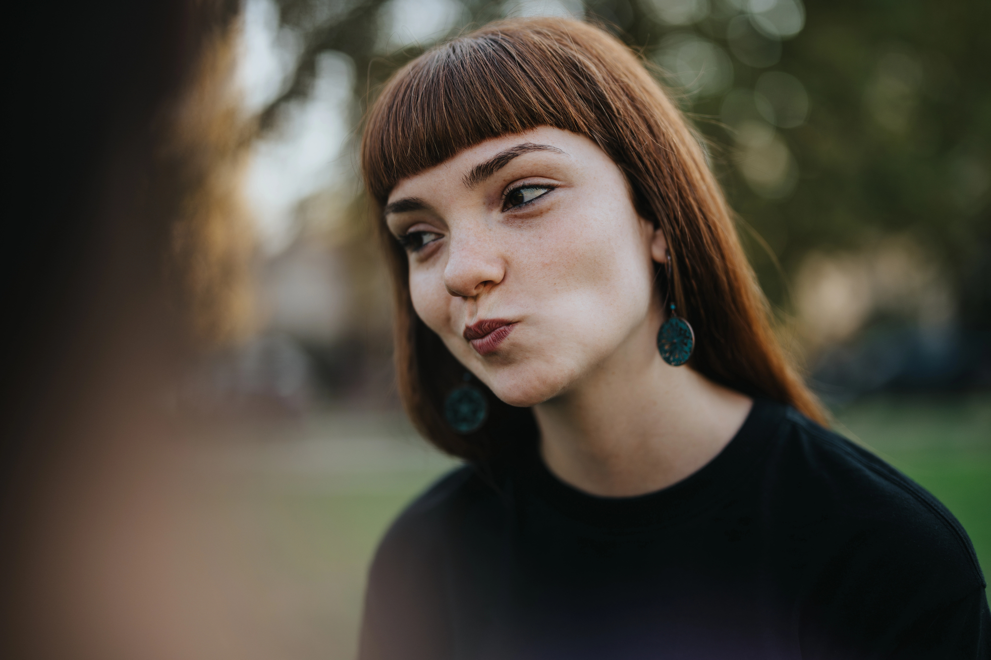 A woman with long red hair and bangs is outdoors, wearing turquoise earrings and a black top. She has a playful expression, with slightly puckered lips. The background is blurred greenery.