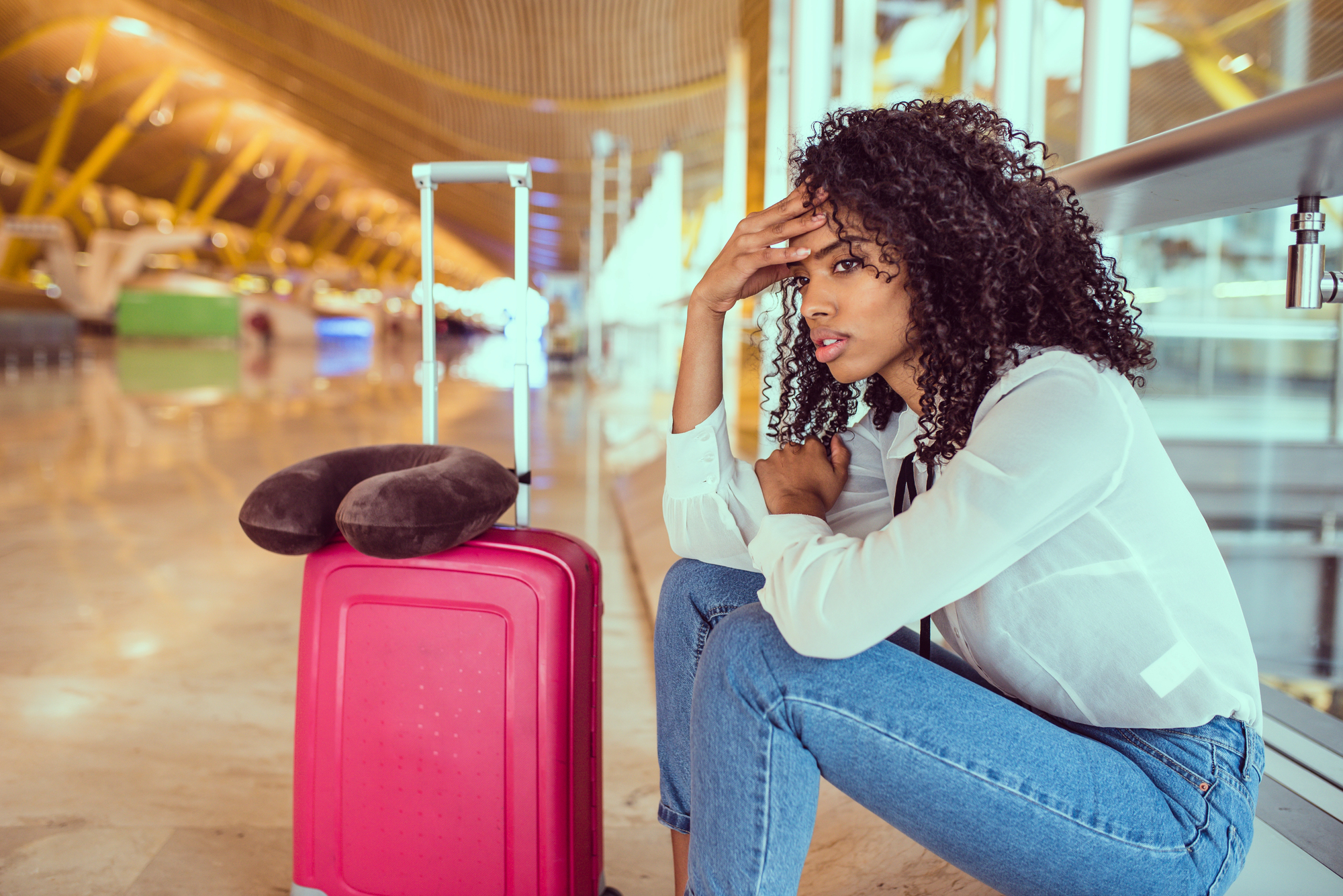 A woman with curly hair sits on the floor of an airport, looking pensive. She rests her head on her hand next to a pink suitcase with a neck pillow on top. Bright lights and airport benches are visible in the background.