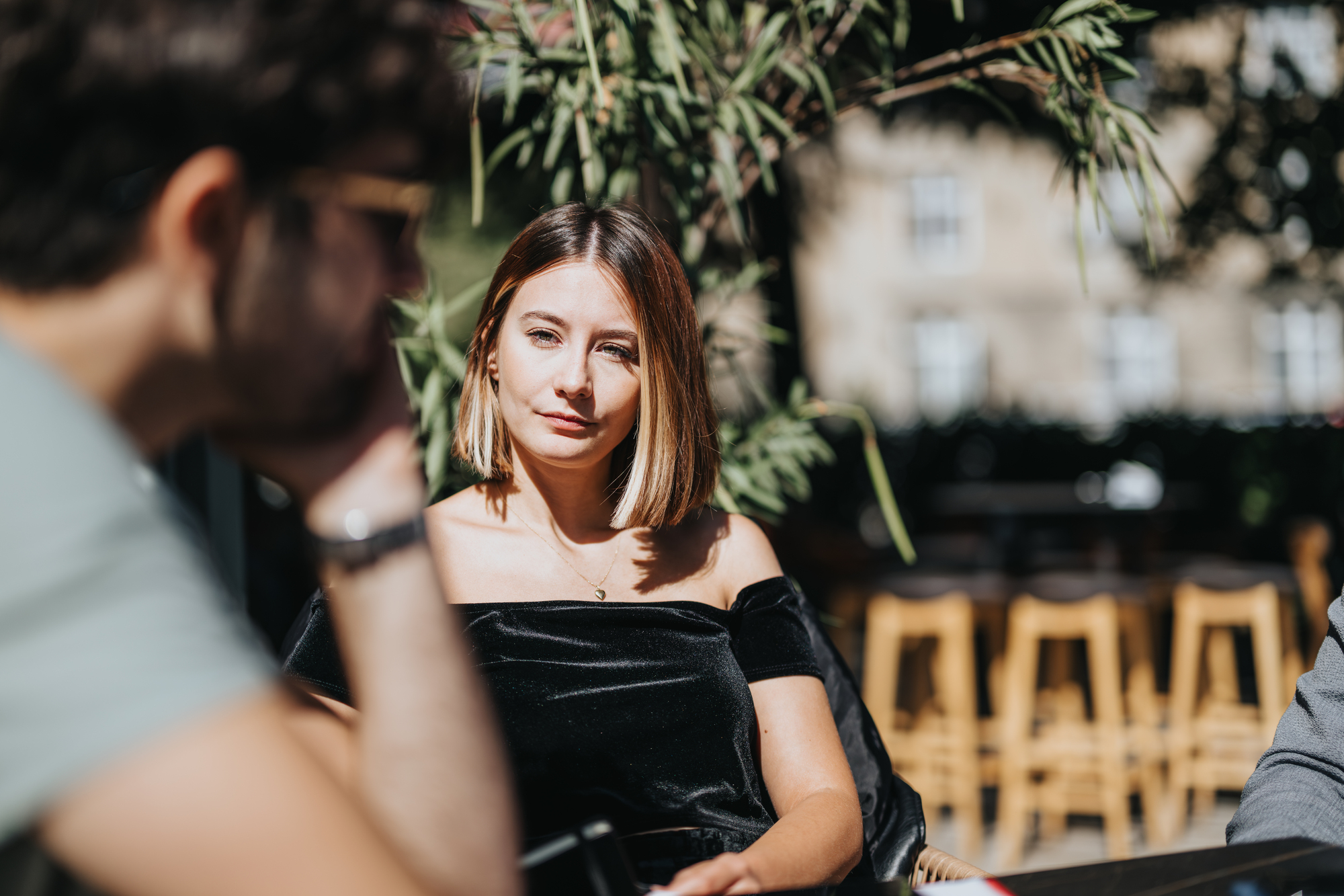 A woman with shoulder-length hair and an off-the-shoulder top sits outdoors, looking at someone out of focus in the foreground. Sunlight filters through leaves above her, casting shadows. Background shows empty bar stools and a building.