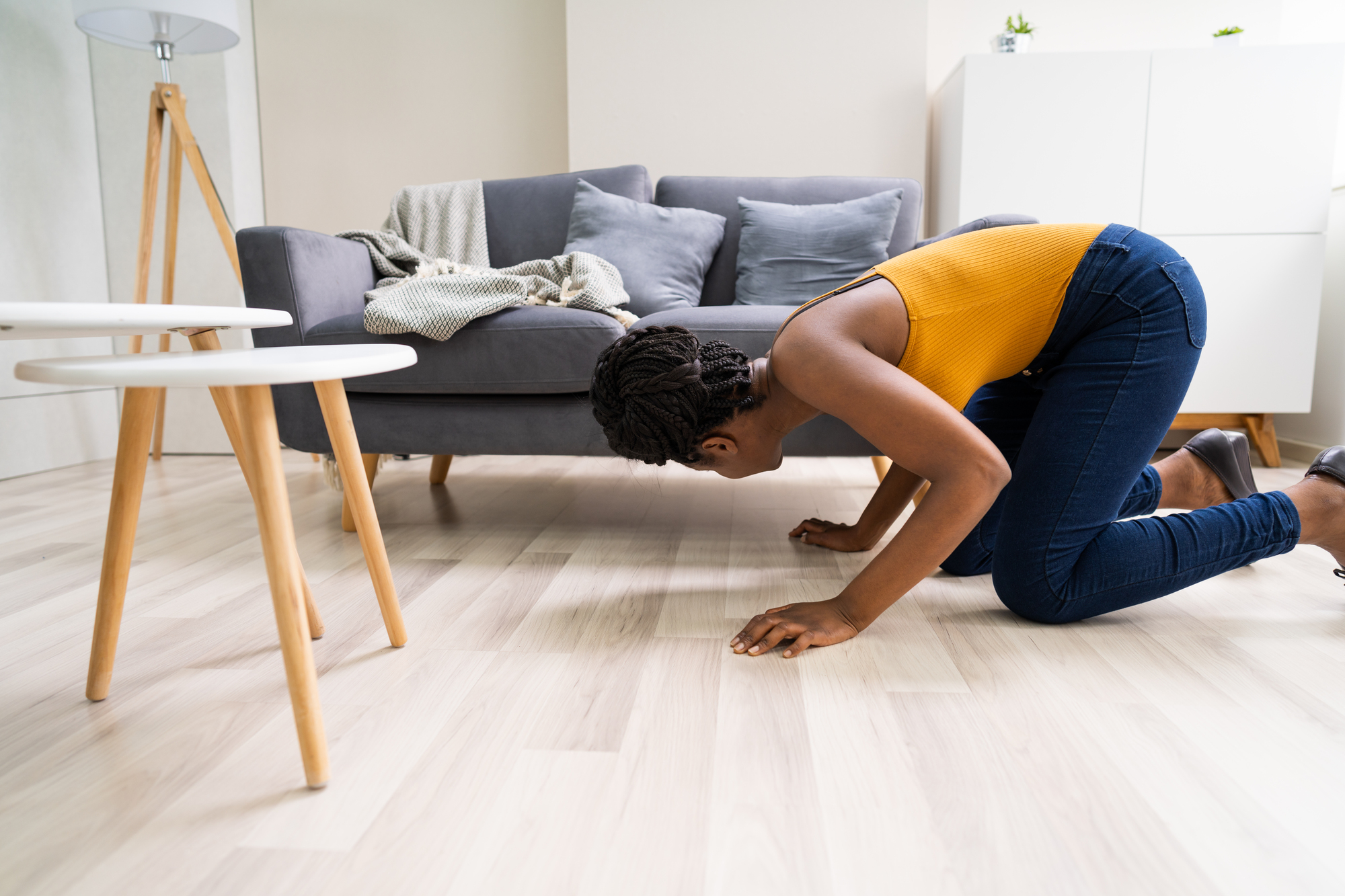 A person kneels on a light-colored wooden floor, peering under a gray sofa. They are wearing a yellow top and blue jeans. Two small round tables are in the foreground. The room has a modern and minimalist design.