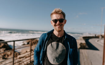 Smiling man with sunglasses and a beard, wearing a dark t-shirt and denim shirt, stands on a boardwalk by the ocean. The background shows waves, rocks, and a clear blue sky.